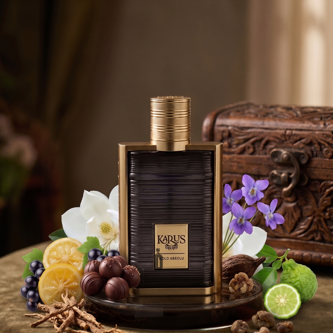 A bottle of Karus Gold Absolu 100ml EDP by Khadlaj, a luxurious fragrance, is surrounded by lemons, chocolates, flowers, grapes, lime, and herbs, with a decorative wooden chest in the background on a green fabric surface.