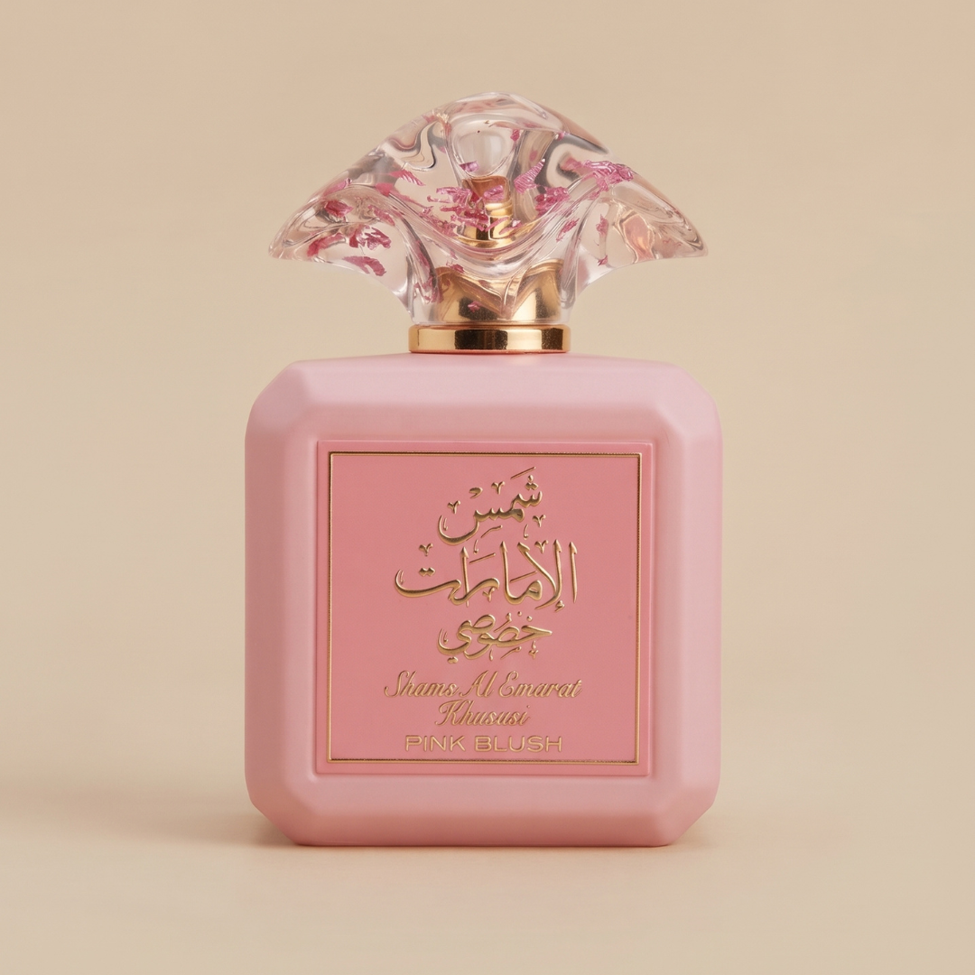 A pink perfume bottle featuring a decorative clear cap, gold Arabic and English text, and “Pink Blush” displayed on the front, set against a beige background—this elegant women’s fragrance is Ard Al Zaafaran’s Shams Al Emarat Khususi Pink Blush 100ml EDP.