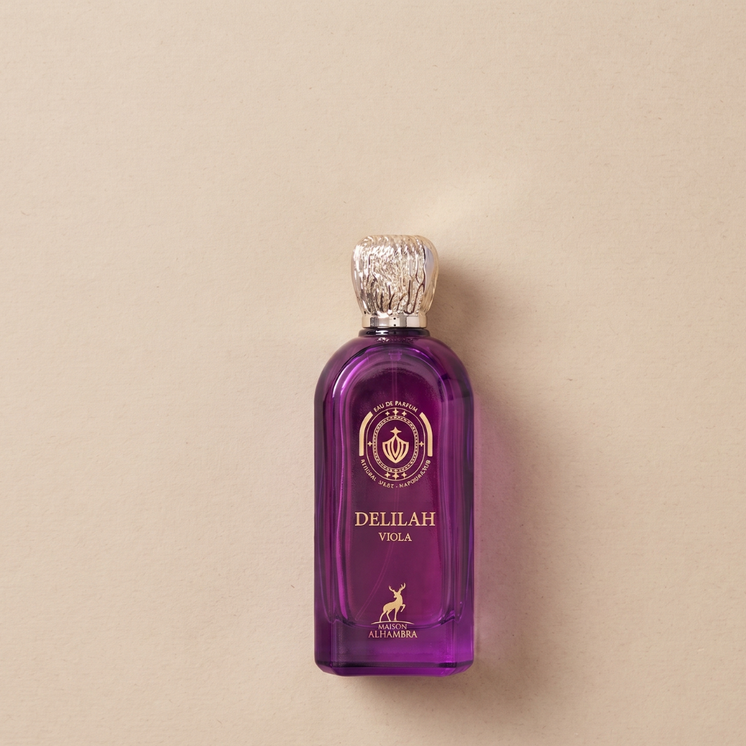 A luxurious 100ml Eau de Parfum, Delilah Viola by Maison Alhambra, features a striking purple bottle adorned with gold accents and a textured silver cap. Presented against a beige background, it exudes sophisticated elegance and alludes to the enchanting floral notes that define this captivating fragrance.