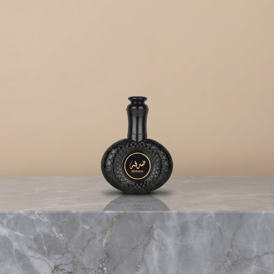 A black, textured perfume bottle adorned with gold Arabic script and the “SHUOYA” text rests on a gray marble surface against a beige background, capturing the luxurious essence of Ard Al Zaafaran’s Sudfa 100ml Eau de Parfum and hinting at its rich coffee-inspired fragrance.