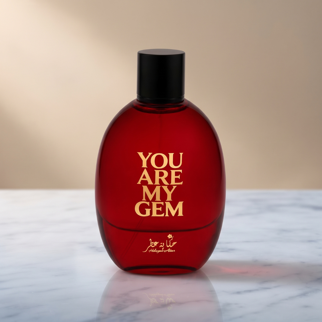 A luxurious red bottle of You Are My Gem 100ml EDP by Hekayat Attar, topped with a sleek black cap, rests elegantly on a marble surface. Gold lettering spells out “YOU ARE MY GEM” on the front, accompanied by finer gold text below. Evoking the allure of a fruity gourmand fragrance, the scene exudes sweetness and sophistication against a softly blurred beige background.