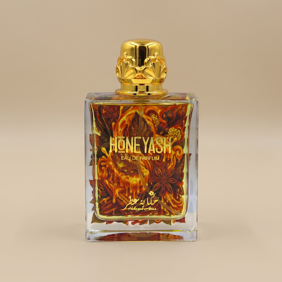 A rectangular bottle of Hekayat Attar’s HoneyAsh 100ml Eau De Parfum stands out with its gold cap and intricate fiery floral motif adorning the front, set against a soft beige backdrop. This exquisite fragrance by Hekayat Attar showcases the inviting warmth of an oriental honey gourmand blend, beautifully reflected in its sophisticated design.