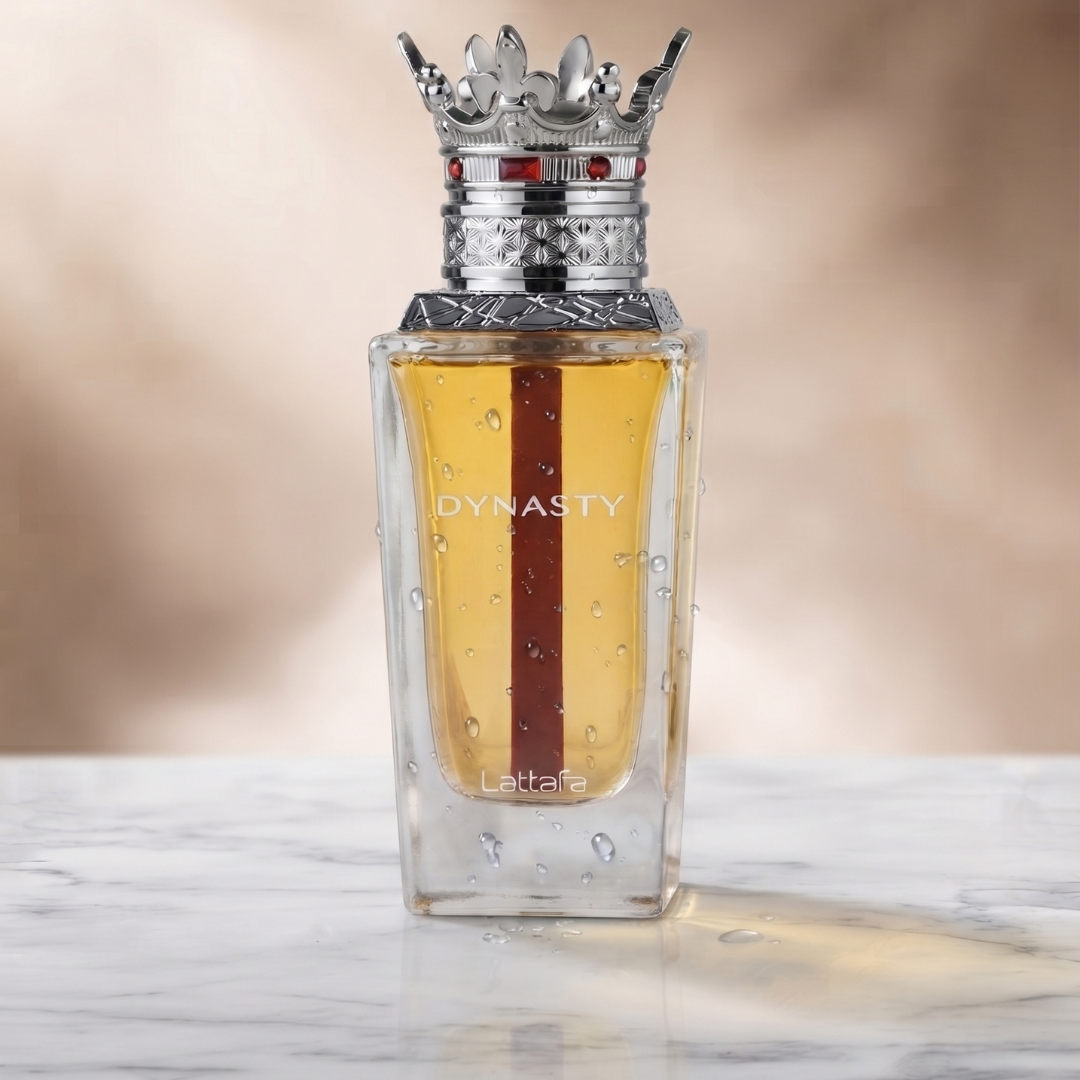 A glass perfume bottle labeled “DYNASTY,” topped with a silver crown-shaped cap, rests on a marble surface adorned with water droplets and displays a brown stripe inside—the elegant design of Dynasty 100ml EDP by Lattafa, a men’s fragrance exuding refined sophistication and the smooth allure of suede.