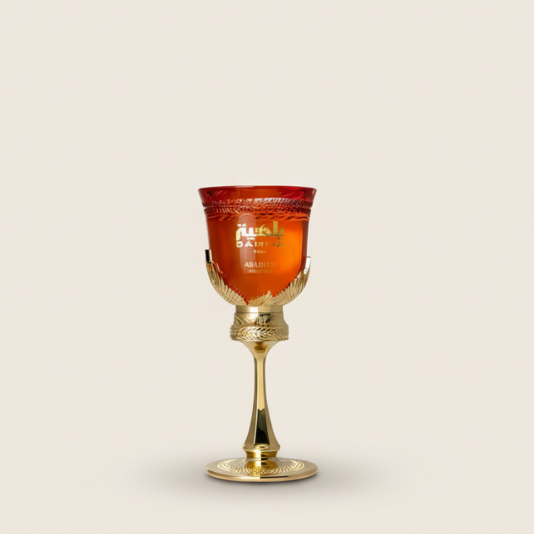 The Bahiya Ruby 100ml EDP by Arabiyat Prestige, a red glass goblet with gold accents on an ornate gold stem and base, evokes a sensual allure against a plain light beige background.