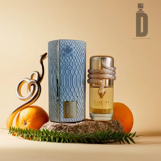 A bottle of Musamam White Intense 100ml EDP by Lattafa, adorned with coiled rope, stands on a stone beside a blue snakeskin-patterned case. The setup is tastefully decorated with oranges, green leaves, and a gold snake decor, displaying the fragrance notes of this sophisticated perfume against a beige background.