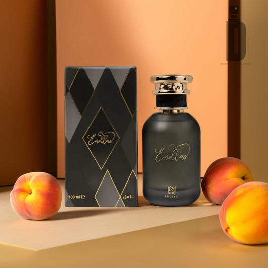 A photograph of a perfume bottle labeled "Endless 100ml EDP - Ahmed Al Maghribi" alongside its packaging box, both featuring a sleek black and gold design. Three peaches are positioned around the bottle and box on a beige surface, with a warm, gradient brown background, hinting at the sophisticated scent of this Eau de Parfum from Ahmed Al Maghribi.