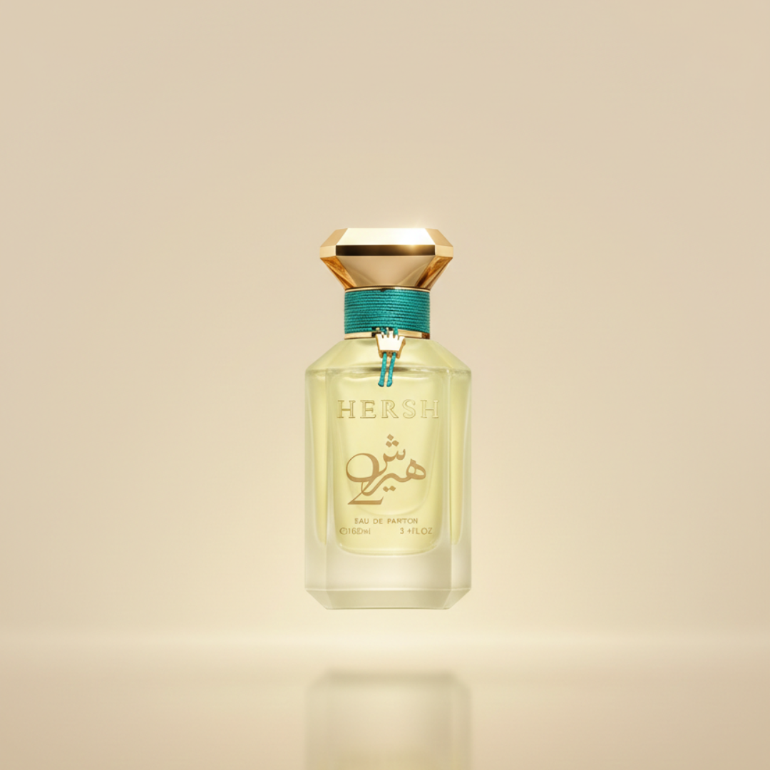 A clear perfume bottle labeled "HERSH" with gold and teal accents stands against a soft beige background. The geometric gold cap tops Hersh 2 - Alezz Oud by Alezz Oud, a vibrant citrus spice fragrance for any occasion.