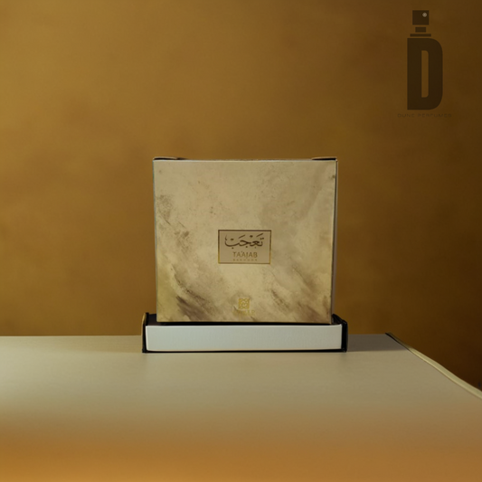 A square box of Ta'ajab Bakhoor (6 tablets) by Ahmed Al Maghribi, featuring a marble-like design on a white and wood-colored base, is displayed on a table. The box is adorned with Arabic text and a small gold emblem at the bottom, emitting fragrant notes. The background has a gradient of warm brown tones.