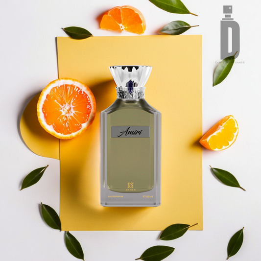 A sleek perfume bottle labeled "Amiri 100ml EDP - Ahmed Al Maghribi" is placed on a yellow sheet surrounded by halved citrus fruits and green leaves, hinting at its luxurious bergamot fragrance. The silver-toned cap adds elegance, while the white background highlights the brand logo "Ahmed Al Maghribi" in the top right corner.