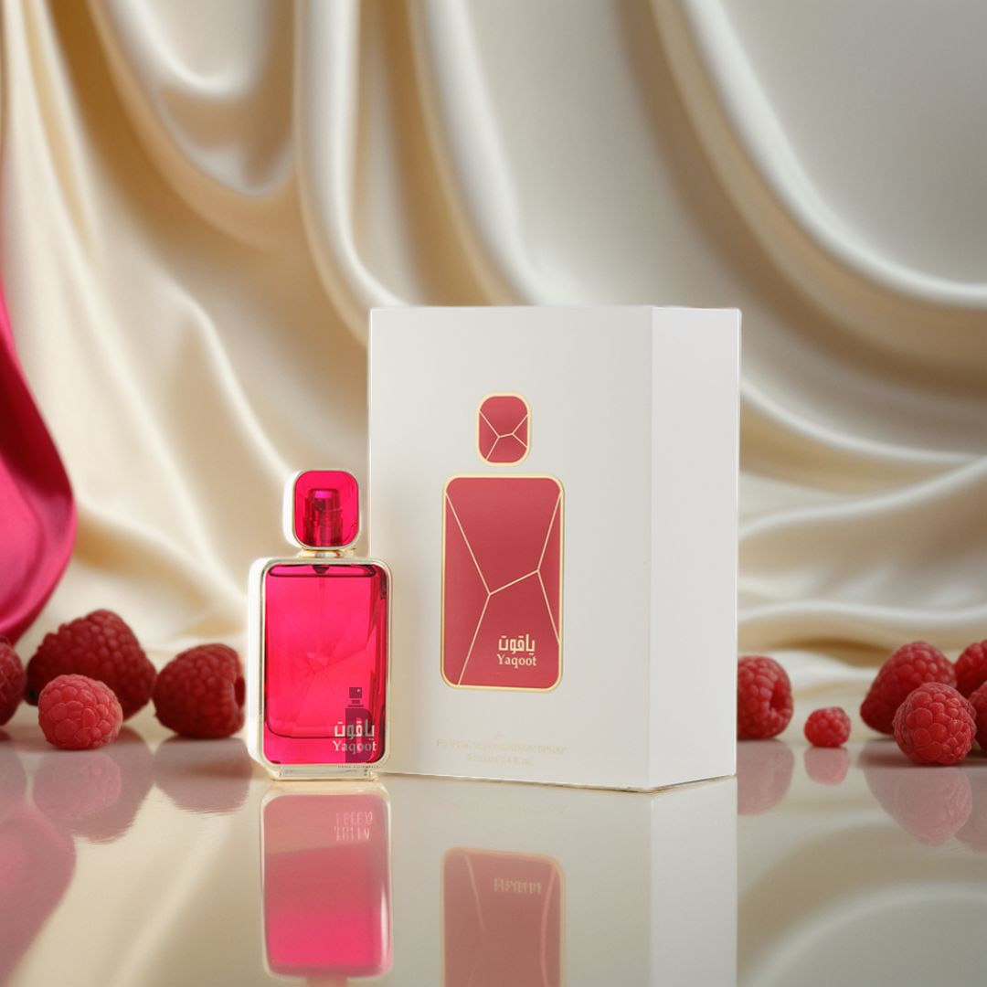 A pink Yaqoot 100ml EDP by Arabian Oud stands beside its white and gold box, with fresh raspberries on a reflective surface and a silky cream fabric backdrop—suggesting the fragrance’s fruity notes.