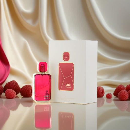 A pink Yaqoot 100ml EDP by Arabian Oud stands beside its white and gold box, with fresh raspberries on a reflective surface and a silky cream fabric backdrop—suggesting the fragrance’s fruity notes.