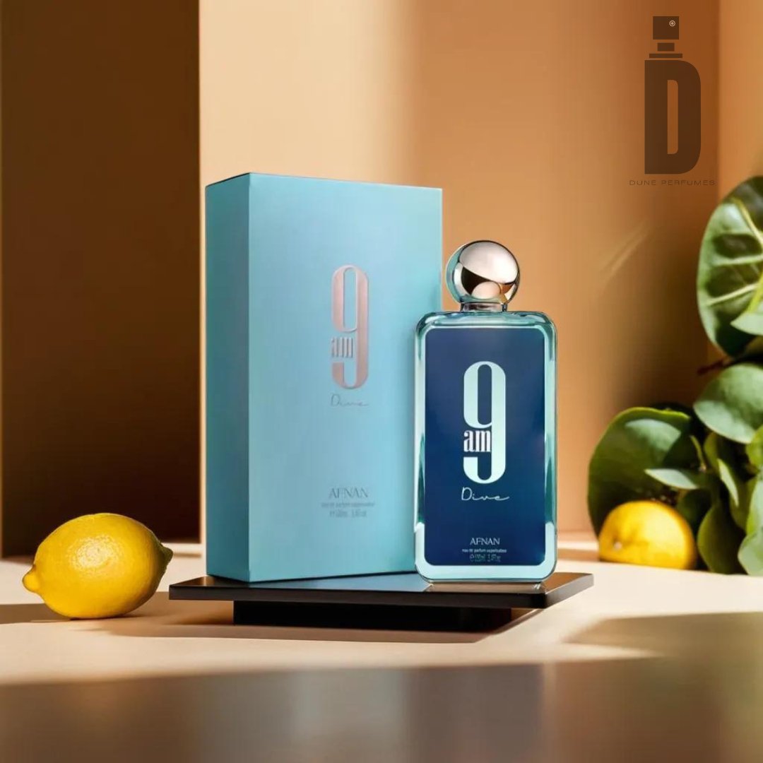 A bottle of 9am Dive 100ml EDP by Afnan, celebrated for its versatile and refreshing fragrance, is displayed next to its matching blue box. Both items rest on a black rectangular tray. Two lemons sit on the table, and green leaves are visible in the background. The scene is brightly lit with a warm tone. The logo "Dunne Perfumery" is in the top right corner.