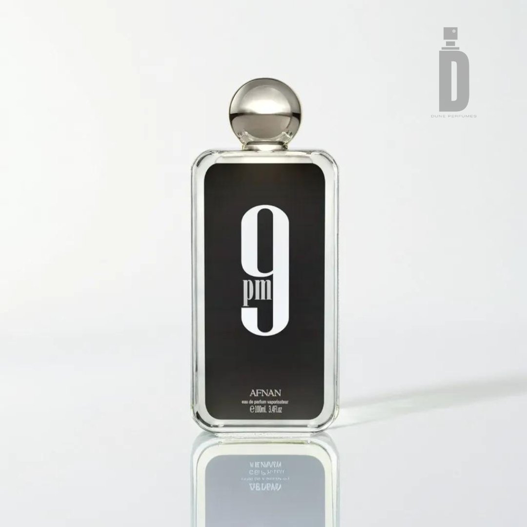 A clear glass bottle of Afnan's "9pm 100ml EDP" perfume, elegantly labeled with bold white text against a black background. The bottle is topped with a silver spherical cap and stands on a sleek, white reflective surface. Hinting at its floral allure, the text reveals the perfume's volume as 100ml (3.4 fl. oz.), promising a truly sensual experience.
