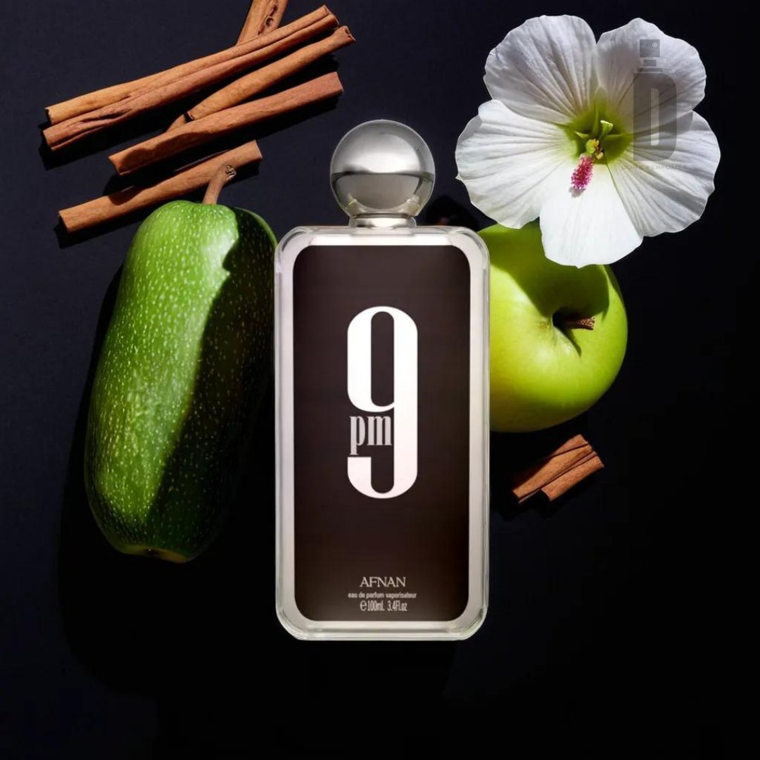 A bottle of "9pm 100ml EDP - Afnan" by Afnan is positioned against a dark background, surrounded by a few pieces of cinnamon sticks, a partly sliced green zucchini, a green apple, and a white flower with a green center.