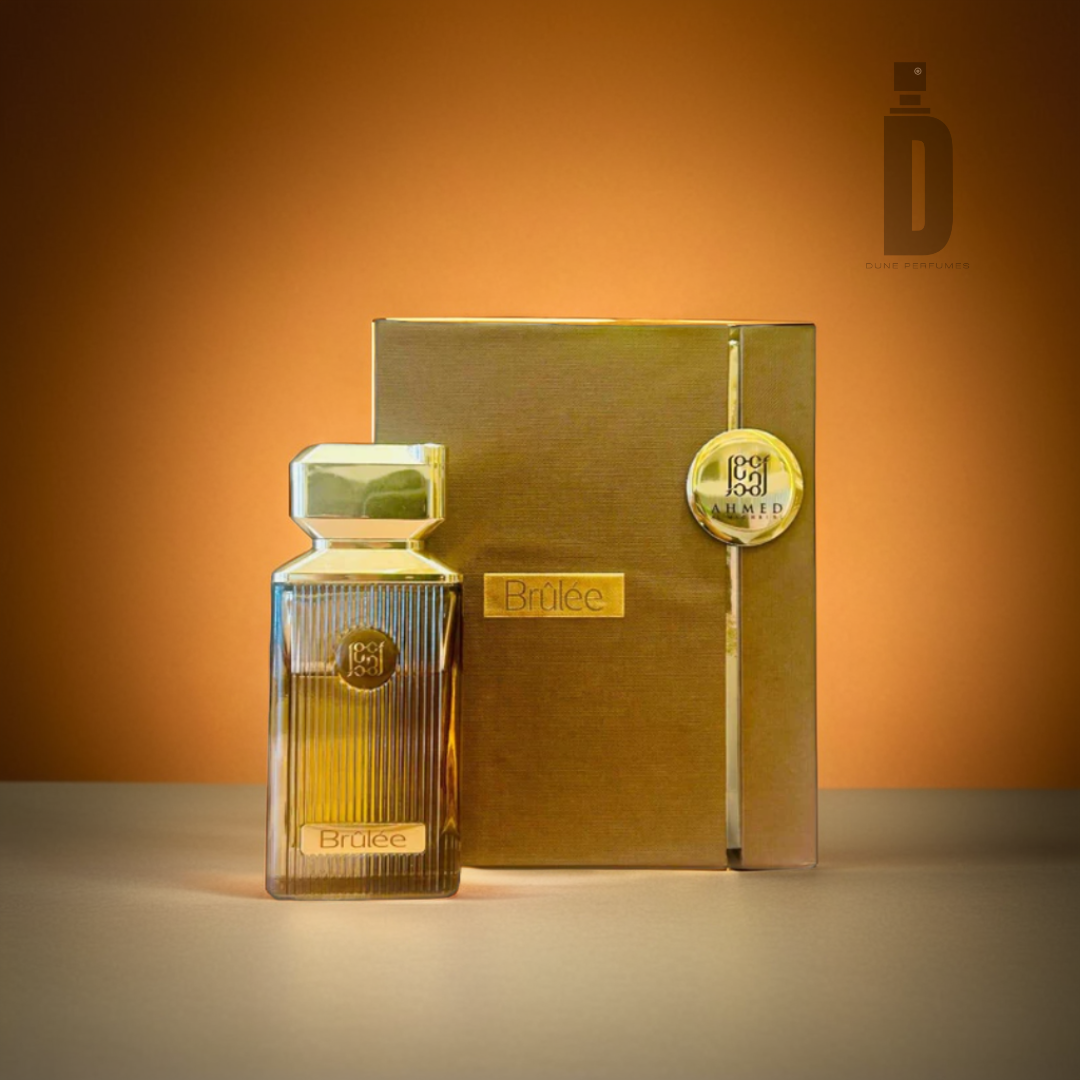 A gold bottle of Brulee 100ml EDP by Ahmed Al Maghribi is placed next to its matching gold rectangular box on a neutral surface. In the background, there's a warm amber gradient with a subtle logo in the top corner.