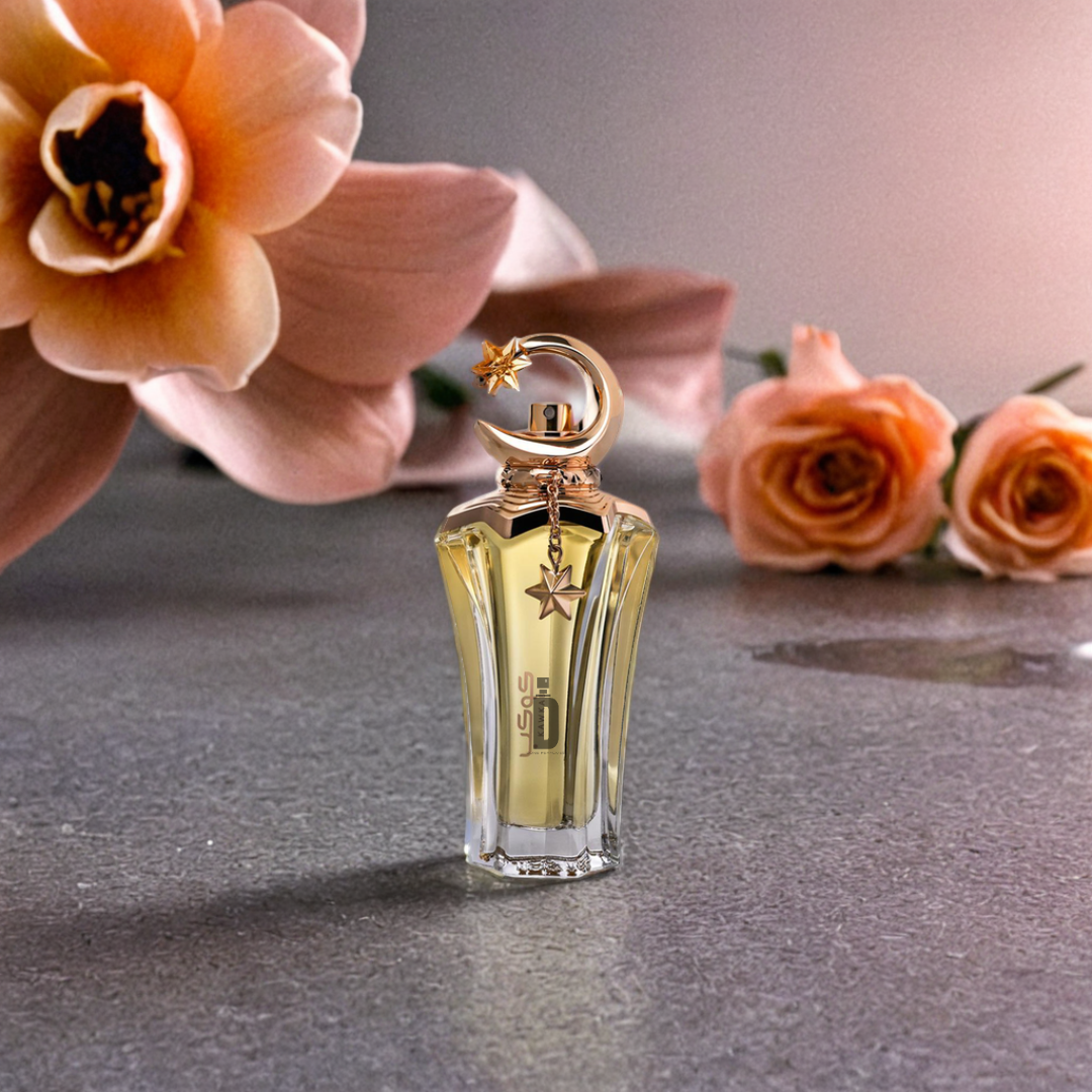 A 100ml Kawkab Eau de Parfum by Ahmed Al Maghribi, in a glass bottle with a gold cap and star charm, rests on a gray surface surrounded by pink and peach flowers—possibly Isparta rose—with petals and water droplets nearby.