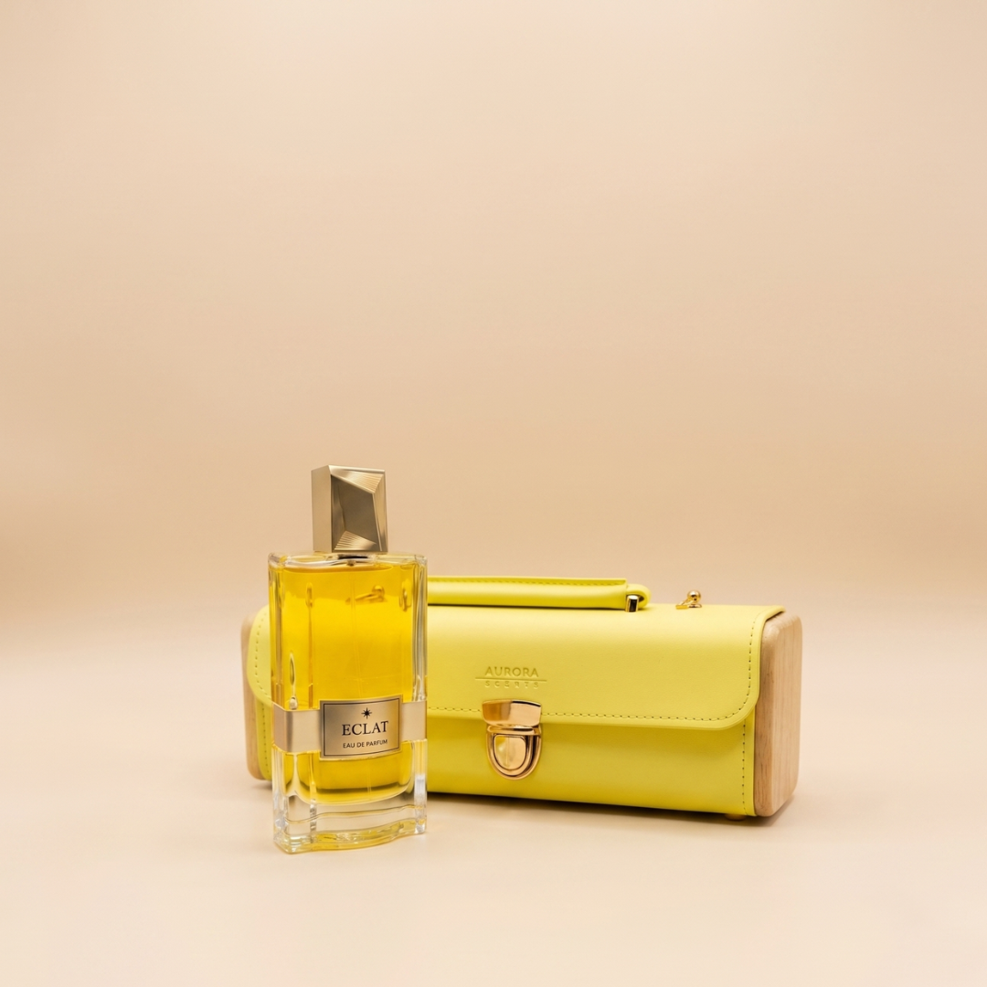 A yellow Aurora Scents Eclat 100ml EDP stands before a matching yellow clutch with a metal clasp, both set against a beige background—ideal for fans of fruity floral fragrances.