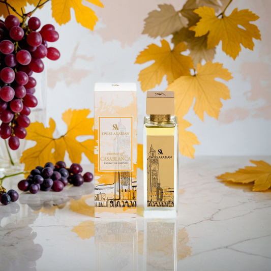 A bottle and box of Swiss Arabian Essence Of Casablanca 100ml EDP are displayed on a reflective surface with artificial yellow leaves and red and black grapes, capturing the fragrance's elegance and the Essence Of Casablanca.