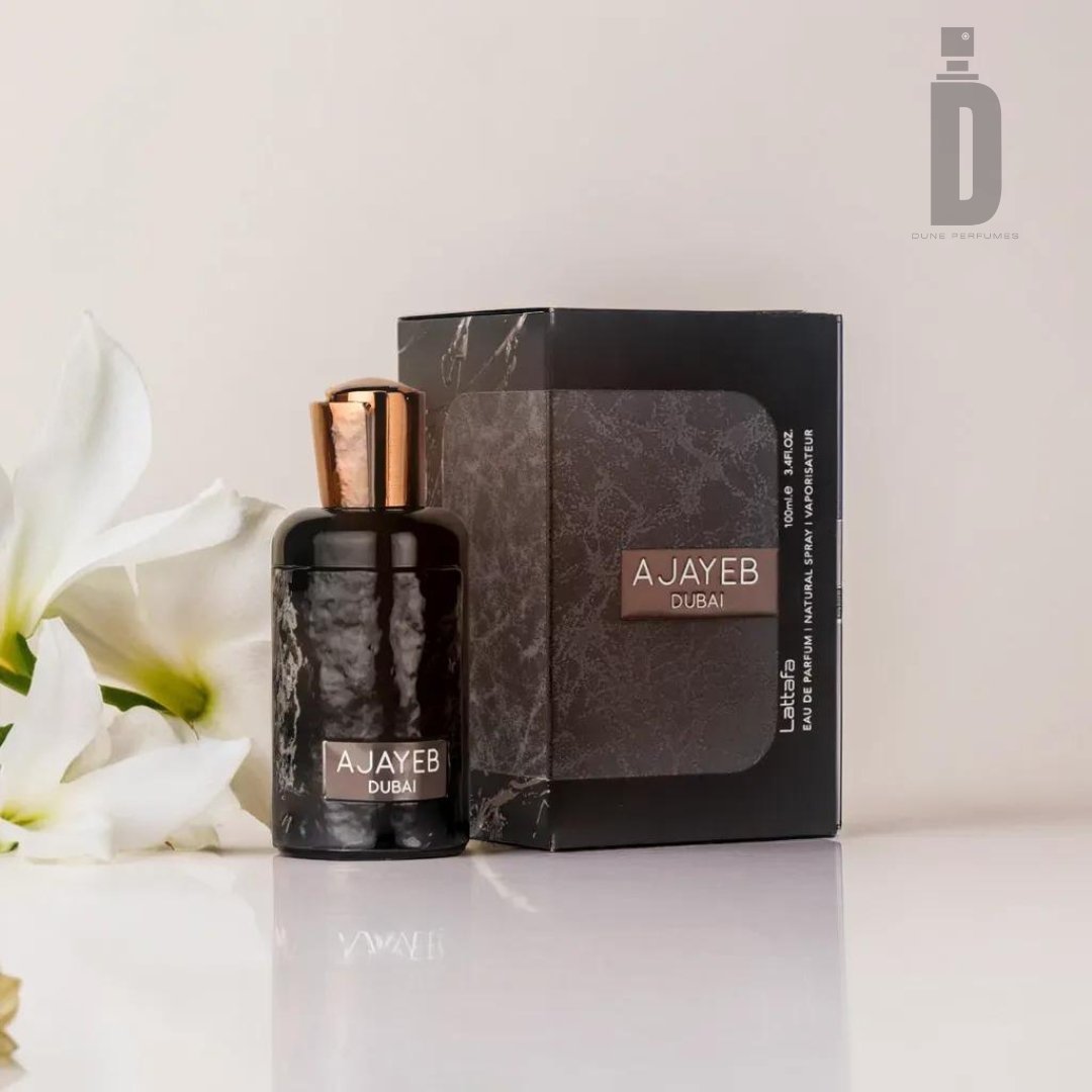 A luxurious black perfume bottle labeled "Ajayeb Dubai 100ml EDP - Lattafa" with a shiny copper cap is placed next to its matching black and copper box. The setup, accented by two white flowers on the left, exudes opulence and mystery. The minimalistic and light background enhances the elegance of this luxury fragrance from Lattafa.