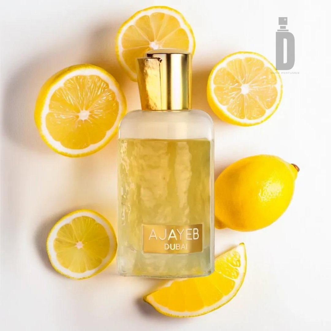 A clear perfume bottle with a golden cap labeled "Ajayeb Dubai Portrait 100ml EDP - Lattafa" is surrounded by halved and whole lemons, arranged on a white surface, highlighting the citrus top notes of this luxurious fragrance from Lattafa.