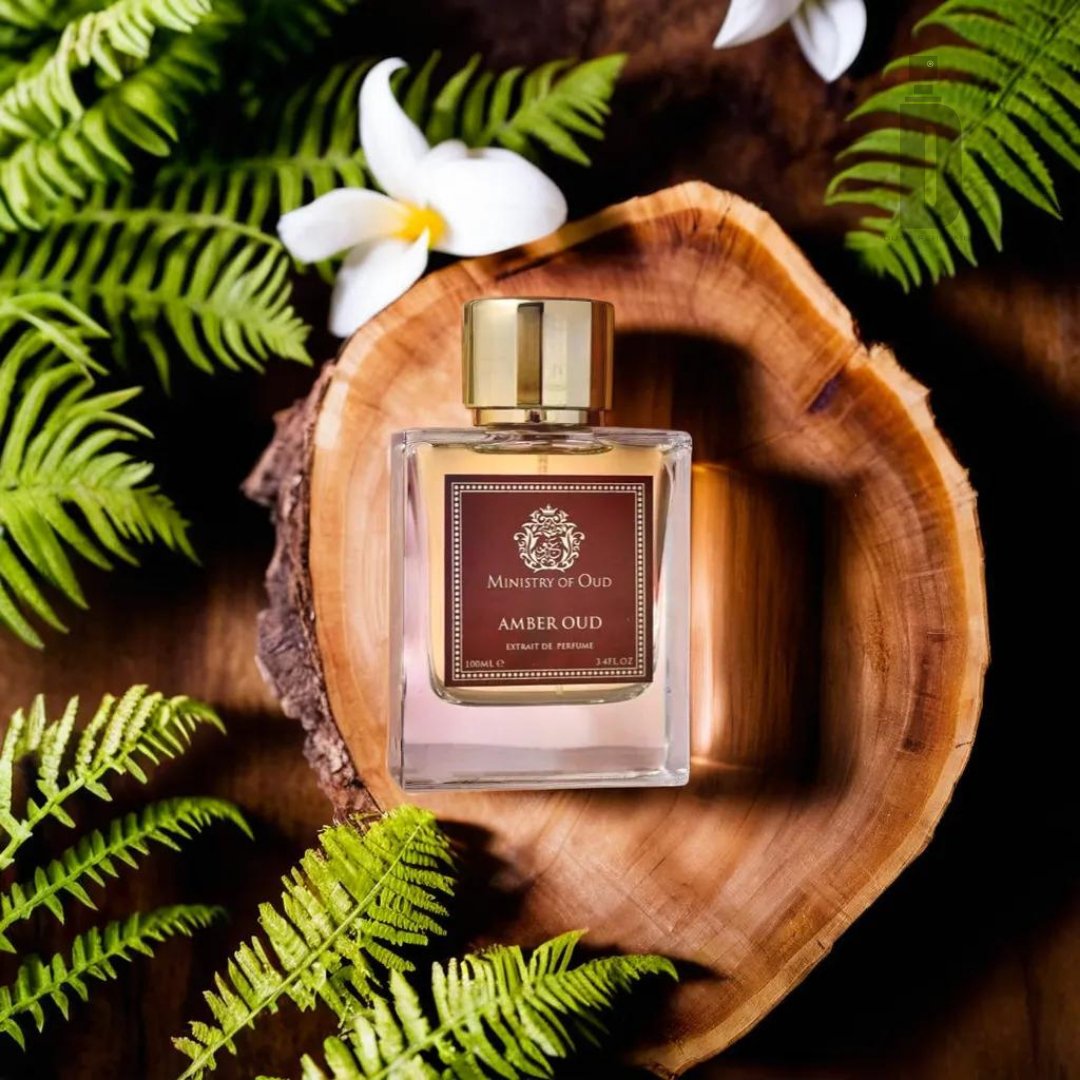 A perfume bottle labeled "Amber Oud 100ml EDP - Ministry Of Oud" from Paris Corner sits inside a rustic wooden bowl surrounded by green fern leaves and white flowers on a wooden surface. The bottle, with its gold cap and elegant design, exudes a timeless allure, perfectly capturing the essence of a luxurious fragrance.