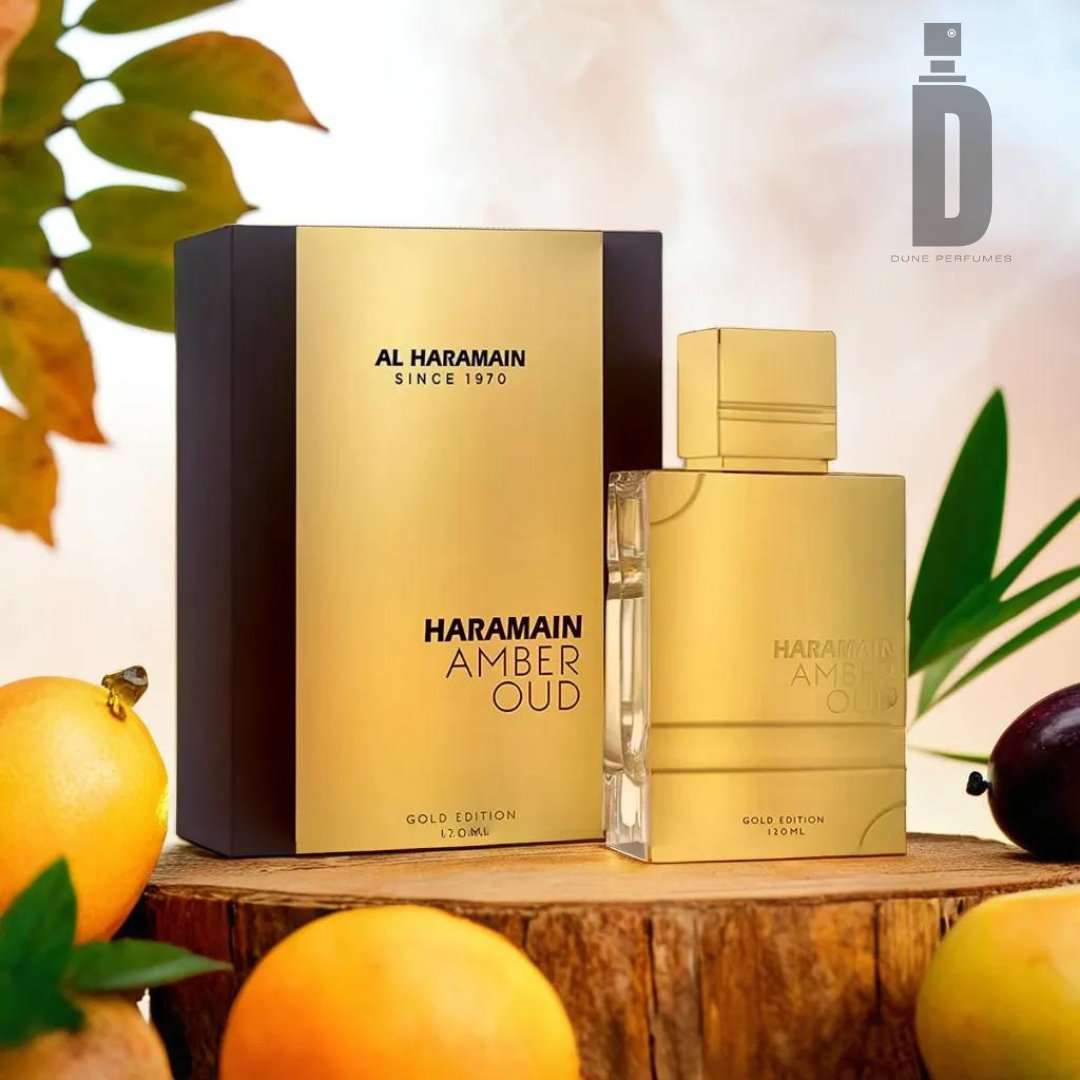 A luxurious golden bottle of Amber Oud Gold Edition 120ml EDP - Al Haramain is displayed alongside its elegant black and gold packaging. The perfume sits on a wooden surface, accompanied by fresh fruits and green leaves in the background, creating a sophisticated ambiance that speaks to its luxurious fragrance with exotic depth.