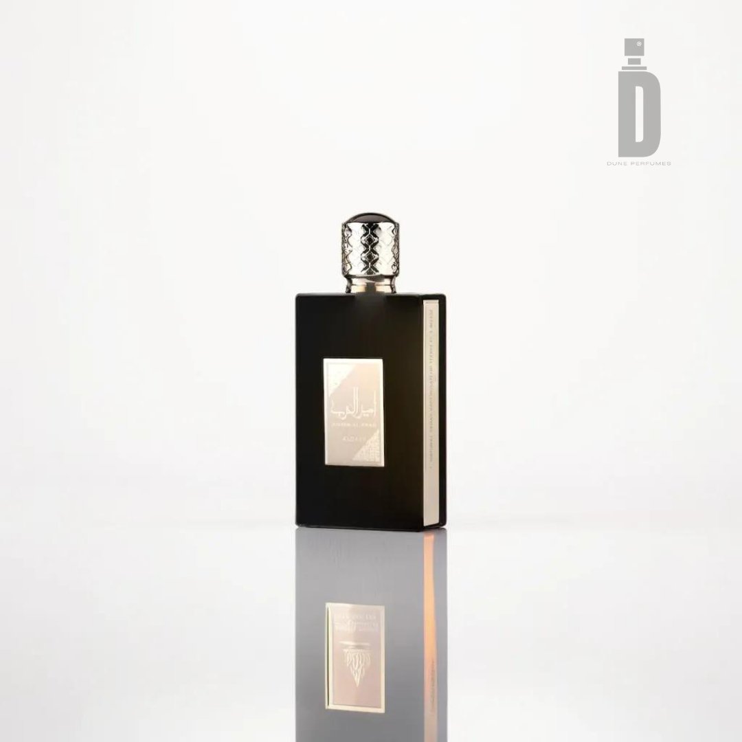 A sleek, square black perfume bottle with a silver cap stands against a plain white background. The bottle has a reflective sheen and an elegant label with Arabic text, exuding regal sophistication. The logo "D" with "Dubai Perfumes" is visible in the upper right corner, heralding the fragrance "Ameer al Arab 100ml EDP - Lattafa" from the renowned brand Lattafa.