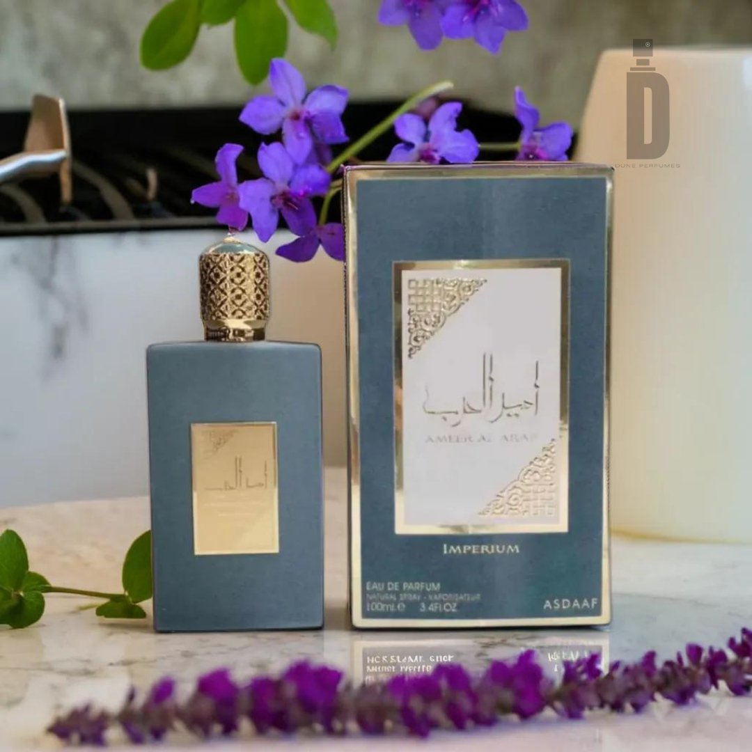 A bottle and box of Ameer Al Arab Imperium 100ml EDP – Asdaaf by Lattafa are displayed on a marble surface. The sleek gray bottle with a gold cap exudes sophistication, while the box features Arabic text and a delicate gold design. Purple flowers and lavender sprigs scattered around create a lasting impression.