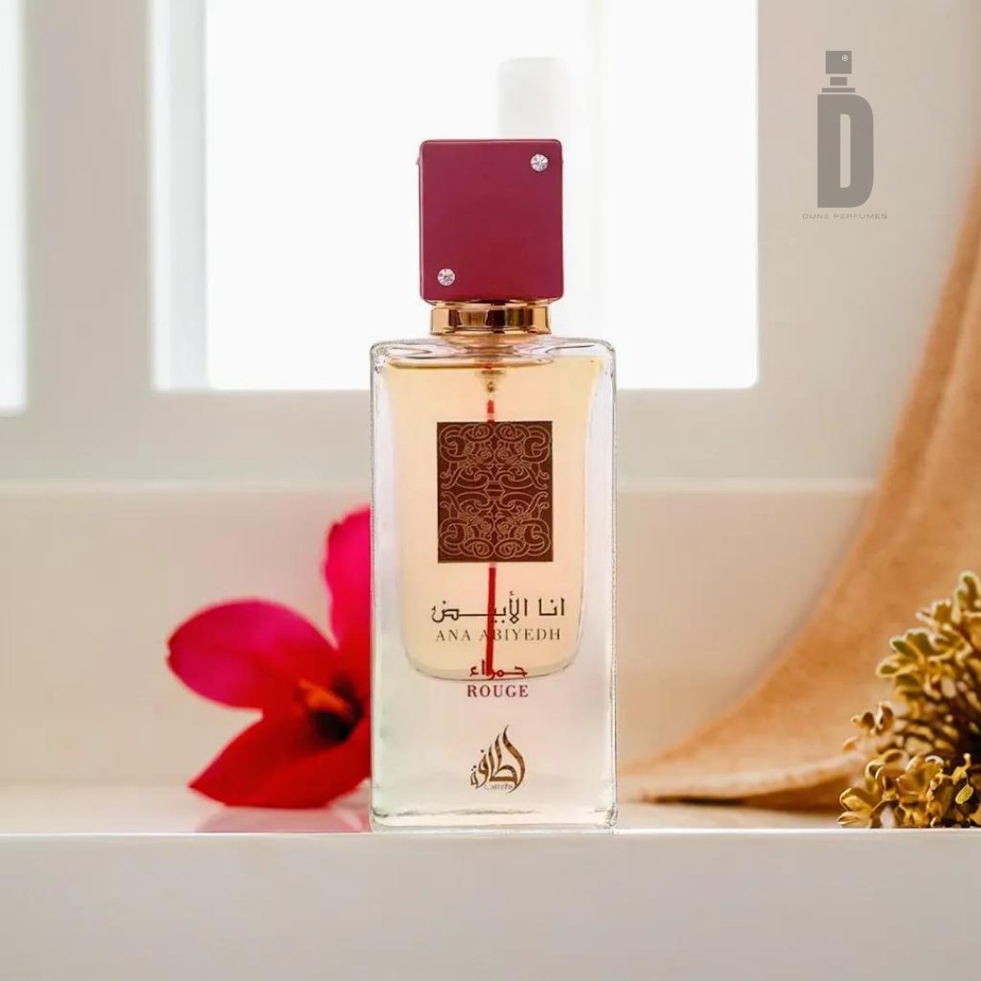 A glass bottle of Ana Abiyedh Rouge 60ml EDP by Lattafa stands on a white surface, backdropped by a blurred, bright red flower and small sprigs with beige buds. The bottle features a red cap and gold accents, with Arabic text and decorative patterns on its front, showcasing the sophisticated fragrance of luxurious ambergris.
