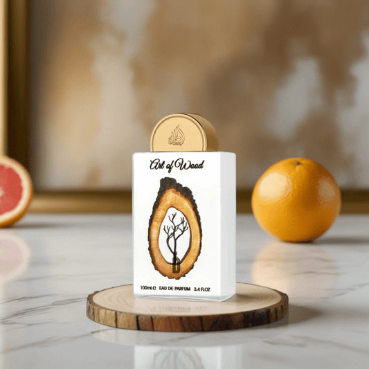 A white luxury "Art Of Wood 100ml EDP - Lattafa" perfume bottle, featuring tree and cedarwood patterns, stands on a wooden coaster with an orange, grapefruit half, and a blurred gold frame for an elegant Lattafa presentation.