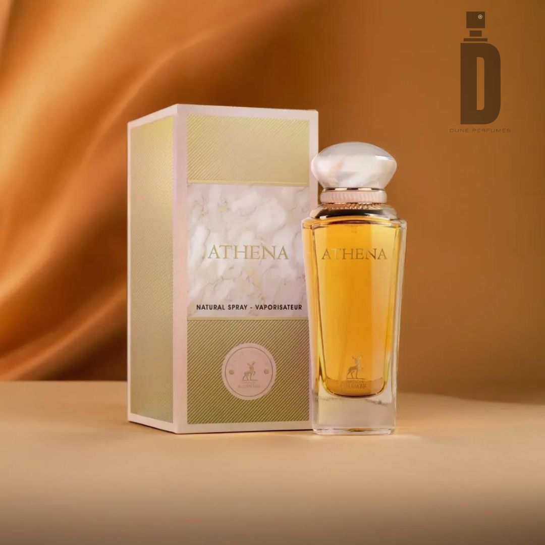 A perfume bottle labeled "Athena 100ml EDP - Maison Alhambra" stands next to its matching, elegant box on a soft, beige surface. The background features a smooth, flowing golden fabric. The box includes the text "Natural Spray - Vaporisateur." The logo of Maison Alhambra is in the top right corner, exuding a divine fragrance.