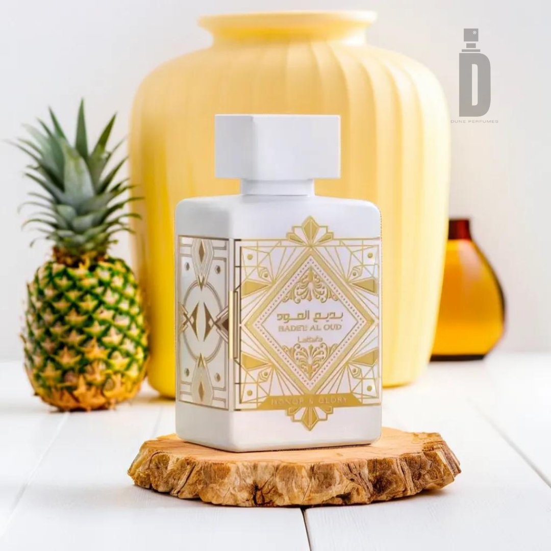 A white, geometric-patterned perfume bottle labeled "Badee Al Oud Honor & Glory 100ml EDP - Lattafa" is displayed on a wooden coaster, exuding an air of valor and magnificence. In the background, a small potted plant, a pineapple, a yellow vase, and a brown vase enhance the scene on a pristine white surface.
