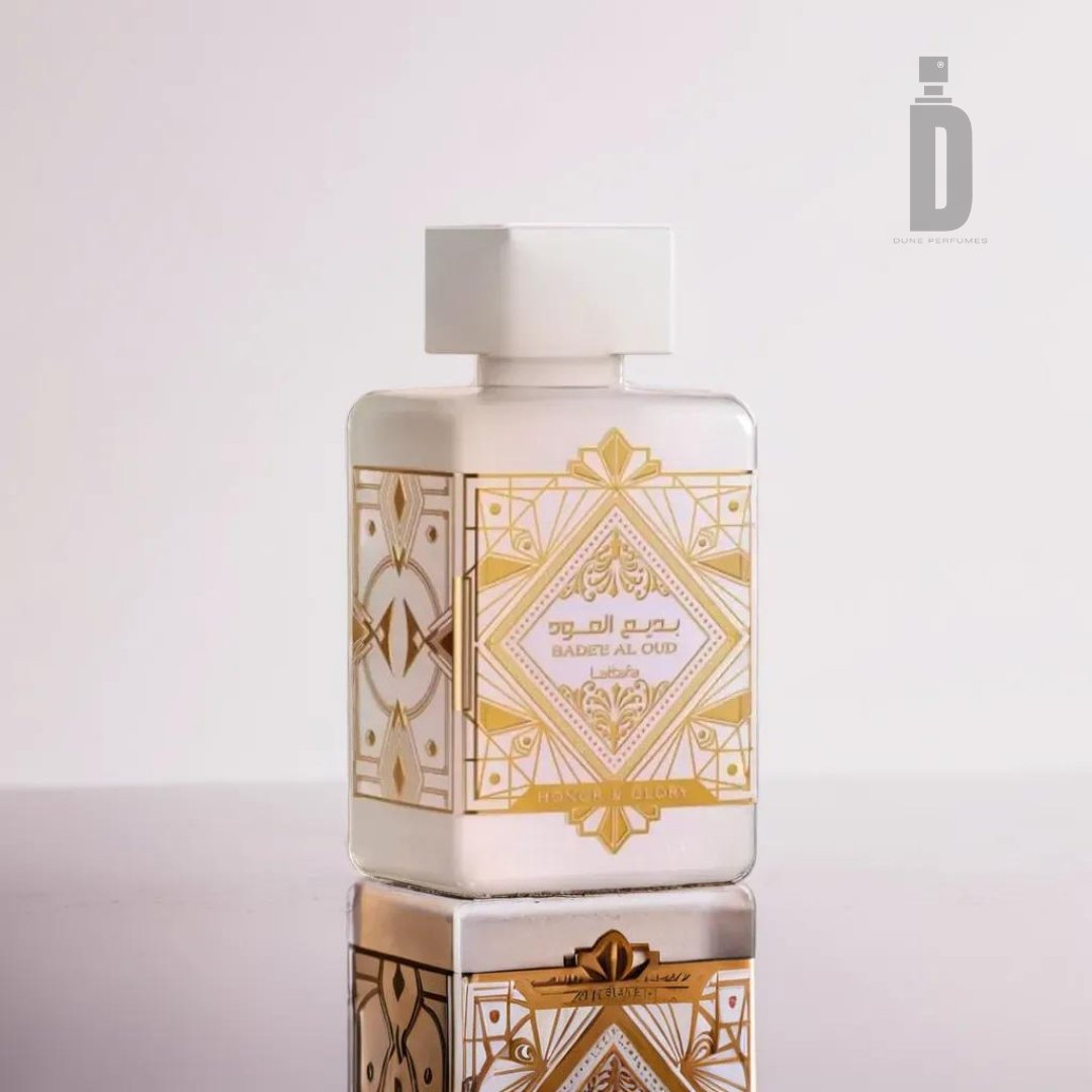 A white rectangular perfume bottle with intricate gold geometric and floral patterns, labeled "Badee Al Oud Honor & Glory 100ml EDP - Lattafa," is displayed on a reflective surface. Exuding valor and magnificence, the luxury fragrance from Lattafa features an elegant "D" logo in the upper right corner against a simple, light gradient background.
