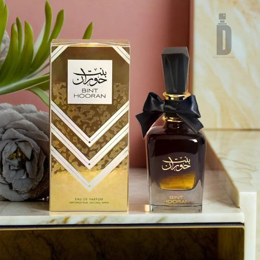 A luxurious perfume bottle labeled "Bint Hooran 100ml EDP - Ard Al Zaafaran" stands next to its matching gold and black box. The bottle, crafted by Ard Al Zaafaran, features a black bow at the neck, a black cap, and a gradient design that exudes timeless sophistication. Both the box and bottle display Arabic text, with a plant accentuating the background.
