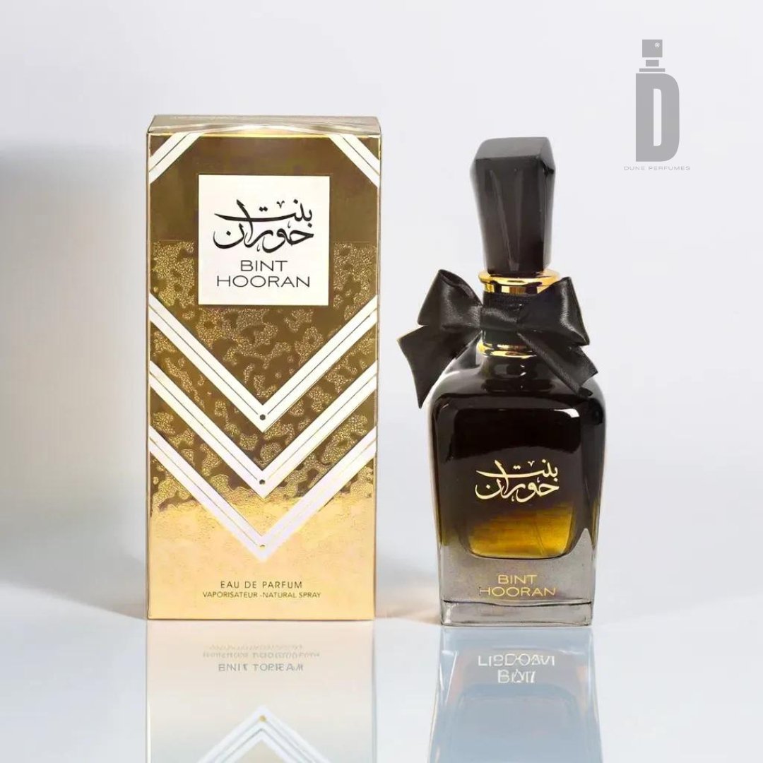 A gold-boxed perfume bottle labeled "Bint Hooran 100ml EDP - Ard Al Zaafaran" exudes timeless sophistication with its matching black-and-gold design. The bottle, adorned with a black bow, sits elegantly next to the box. A logo resembling a stylized perfume bottle graces the upper right corner, inscribed in Arabic and English, highlighting the brand name Ard Al Zaafaran.