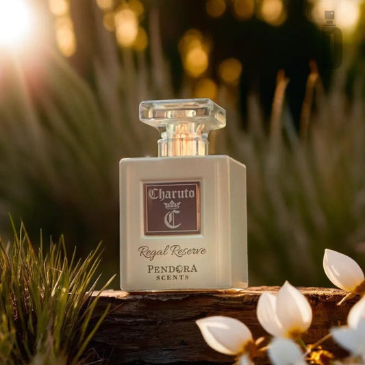 A bottle of Charuto Regal Reserve 100ml EDP by Paris Corner sits on a wooden surface outdoors, surrounded by white flowers and green foliage. Sunlight filters through the background, casting a warm, golden glow, highlighting the sophisticated perfume's elegant glass bottle.
