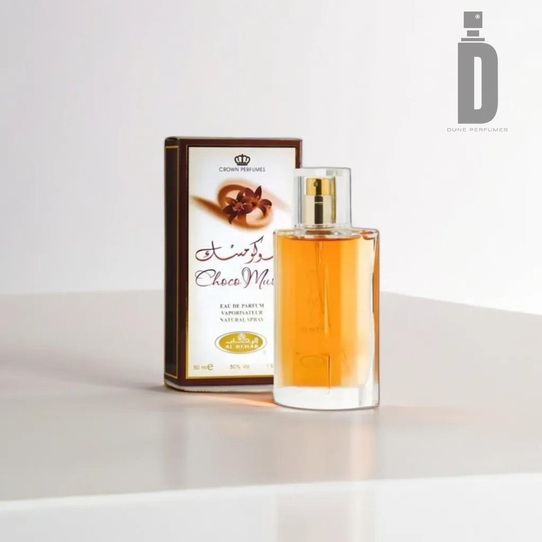 A bottle of Al Rehab's Choco Musk perfume stands in front of its box on a plain surface, radiating a musky scent. The brownish-yellow hue of the perfume complements the box, which features decorative text and an image of chocolate pieces.