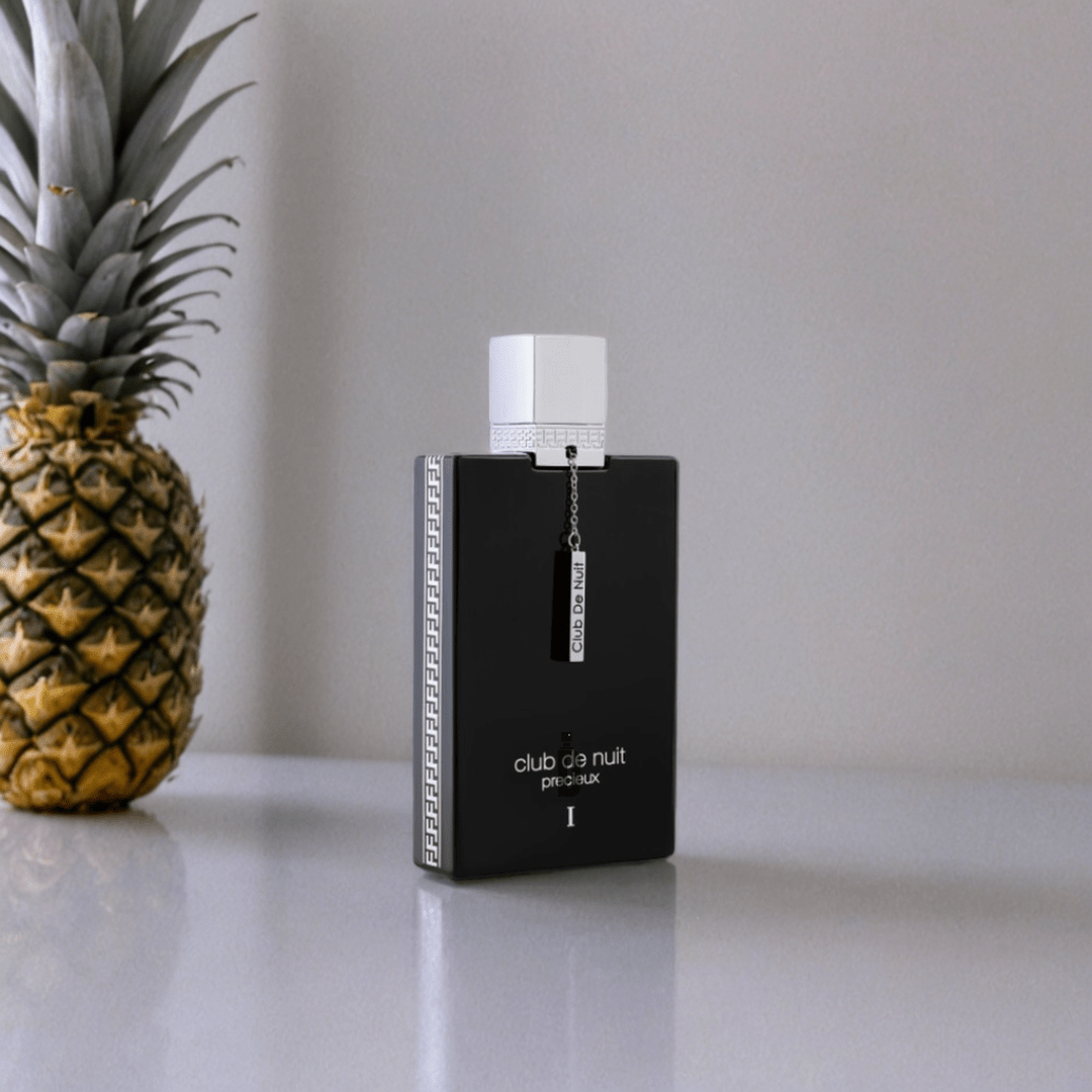 A 55ml bottle of Armaf Club De Nuit Precieux EDP with a white cap sits on a reflective surface beside a pineapple, highlighting this luxurious fragrance against a plain, light-colored background.