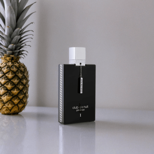 A 55ml bottle of Armaf Club De Nuit Precieux EDP with a white cap sits on a reflective surface beside a pineapple, highlighting this luxurious fragrance against a plain, light-colored background.