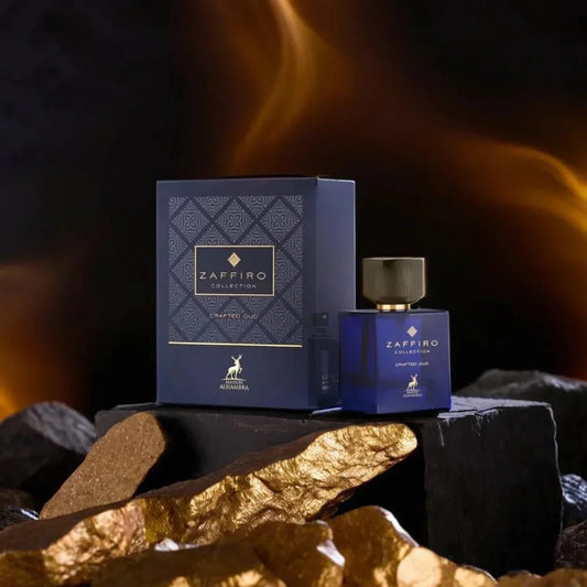 A dark blue perfume box and bottle from the Crafted Oud 100ml EDP - Maison Alhambra collection, presenting a sophisticated fragrance, are displayed among rocks with a golden sheen. The backdrop features an orange and black gradient that evokes fire or intense light. The Maison Al Hambra logo is visible on both the box and bottle.