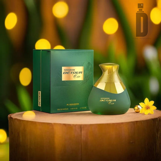 A bottle of Detour Eco 100ml EDP - Al Haramain, a sophisticated unisex fragrance with a woody spicy scent, stands next to its matching green and gold box on a wooden surface. The background features blurred-out warm lights and a small yellow flower, adding a cozy and elegant atmosphere.