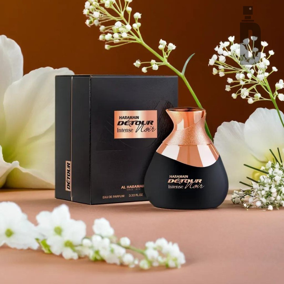 A bottle of Detour Intense Noir 100ml EDP by Al Haramain, featuring a sleek black and copper design, is displayed in front of its box. The presentation is enhanced with delicate white flowers that add an elegant touch. This unisex fragrance is set against an amber-brown background.