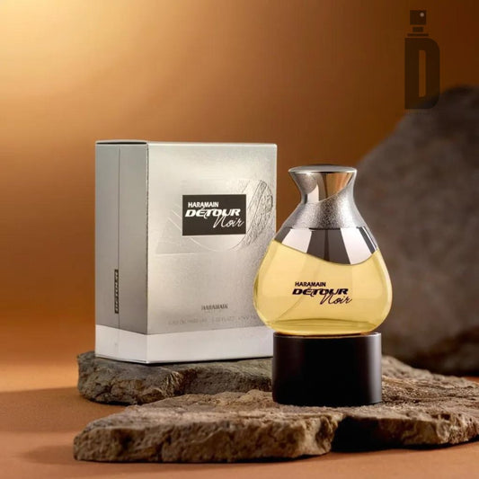 A bottle of Detour Noir 100ml EDP by Al Haramain sits on a textured rock beside its corresponding silver packaging box. The bottle boasts a curvy, modern design with a silver cap and black base, filled with amber-colored liquid—a sophisticated blend exuding luxury fragrance. The background is a warm brown gradient.