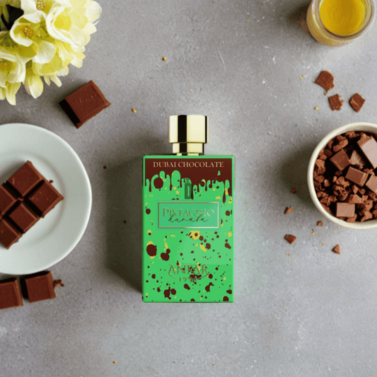 A green bottle of Anfar's Dubai Chocolate Pistachio Kunafa 80ml EDP rests on gray, surrounded by chocolate, crushed chocolate in a bowl, a glass of pale yellow liquid, and yellow flowers—capturing the essence of Middle Eastern desserts.