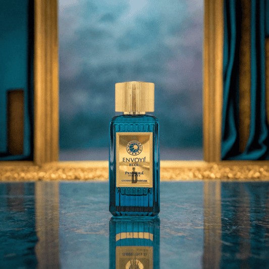 A blue glass bottle of Envoye Blue 100ml EDP by Paris Corner, topped with a gold cap, rests on a reflective marble surface, evoking masculine elegance amid blurred gold-framed windows and blue curtains.