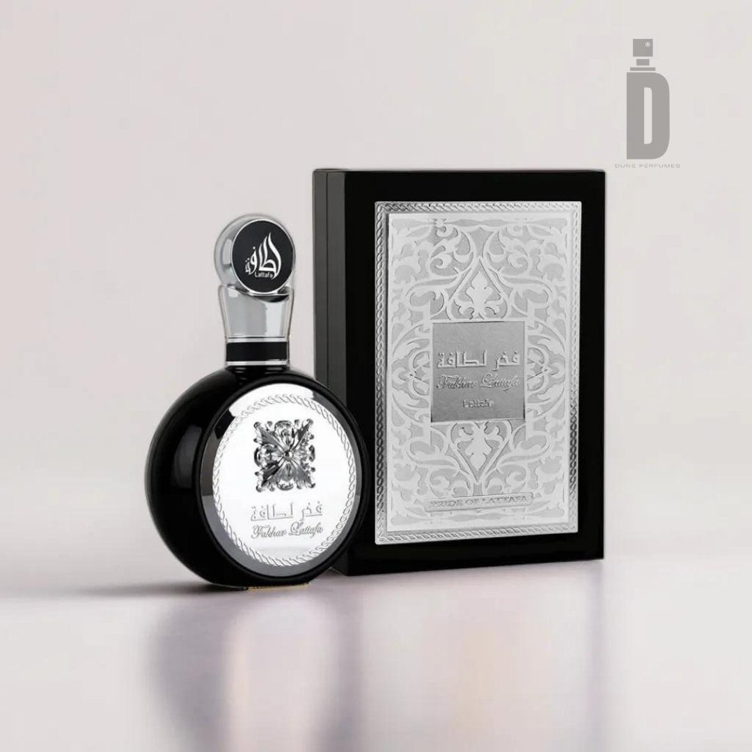 A stylish black perfume bottle with silver detailing, showcasing the sophisticated fragrance of Fakhar Lataffa Black 100ml EDP - Lattafa, is displayed next to its matching elegant box adorned with intricate silver patterns. The bottle's cap features an Arabic inscription, adding a touch of cultural elegance.