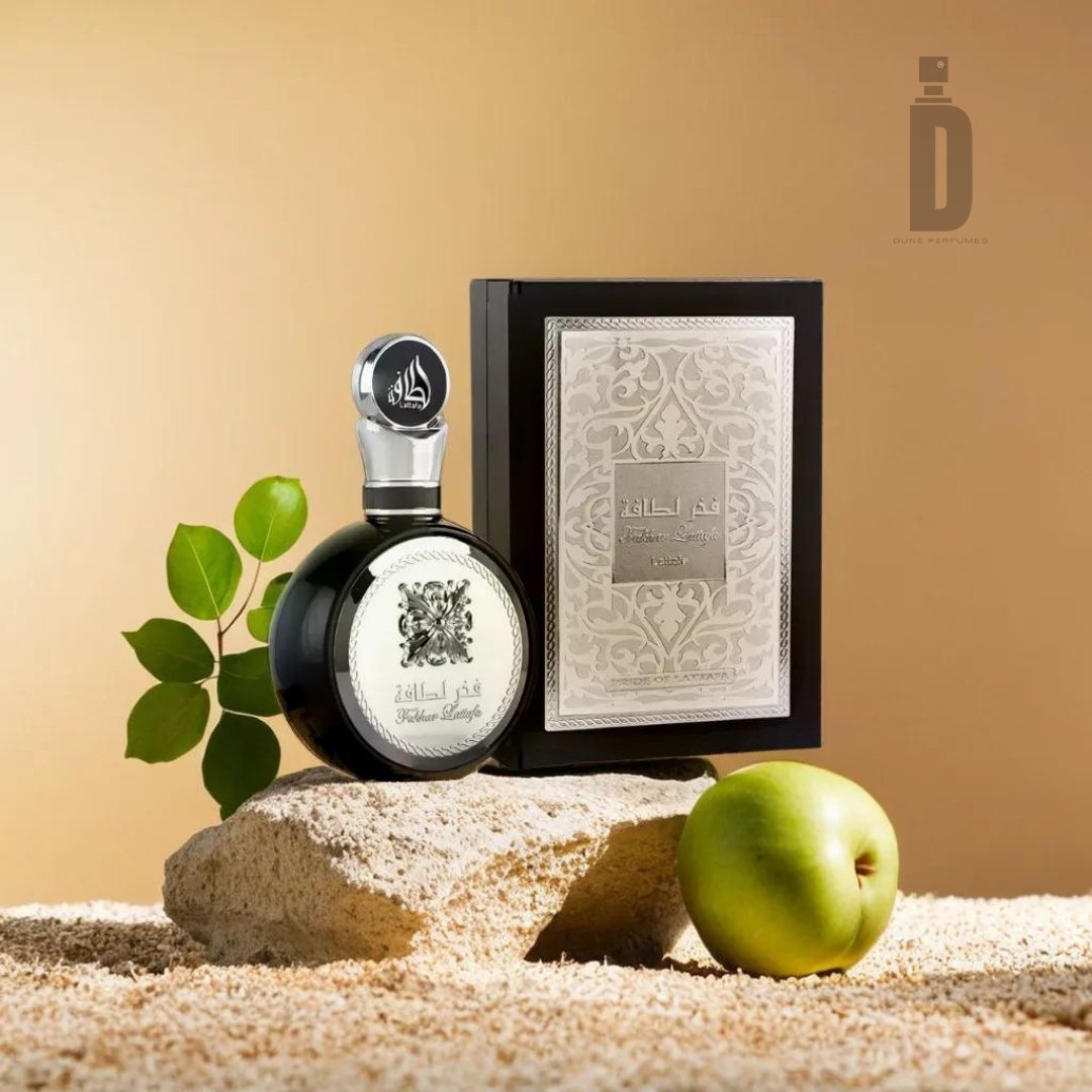 An elegant Fakhar Lataffa Black 100ml EDP - Lattafa perfume bottle with a silver cap and intricate design stands on a stone slab alongside a decorative box. Green leaves and a green apple are placed nearby, all set against a neutral background, giving a luxurious and natural vibe enhanced by its sophisticated fragrance.