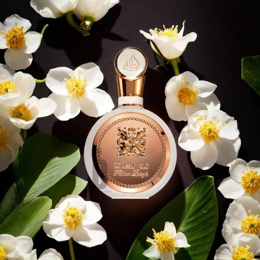 A round, gold-capped perfume bottle of Fakhar Lattafa Rose 100ml EDP is adorned with intricate designs and Arabic script, surrounded by blooming white flowers and green leaves against a dark background. This exquisite perfume by Lattafa radiates a truly feminine fragrance.