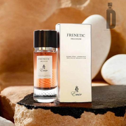 The Frenetic Delicieuse 80ml EDP - Emir by Paris Corner stands next to its matching box on a textured surface with a blurred background. The clear bottle contains a gradient amber liquid that exudes a sweet, warm fragrance, topped with a black and silver cap. The packaging features elegant text and branding.