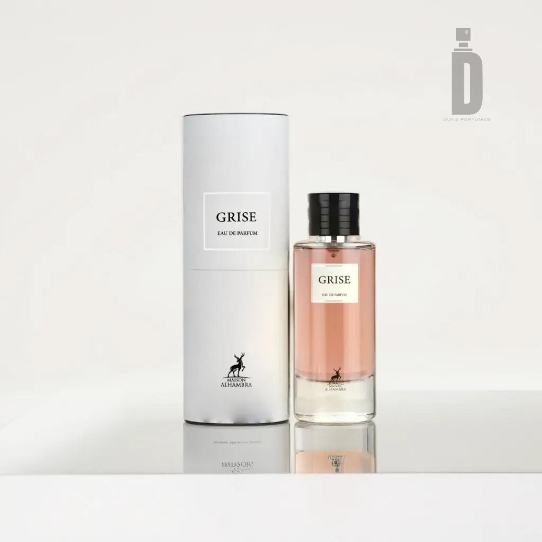 An image showcasing a bottle of "Grise 100ml EDP - Maison Alhambra" next to its cylindrical packaging. The transparent bottle, with a rose-colored liquid inside and a black cap, exudes timeless sophistication. The white packaging with black text features an animal logo, echoing the floral woody notes of the Grise fragrance by Maison Al Hambra.